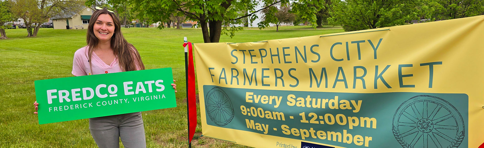 Megan Craggs holds the FredCo Eats sign next to the Stephens City Farmer's Market banner at the grand opening of the new market.